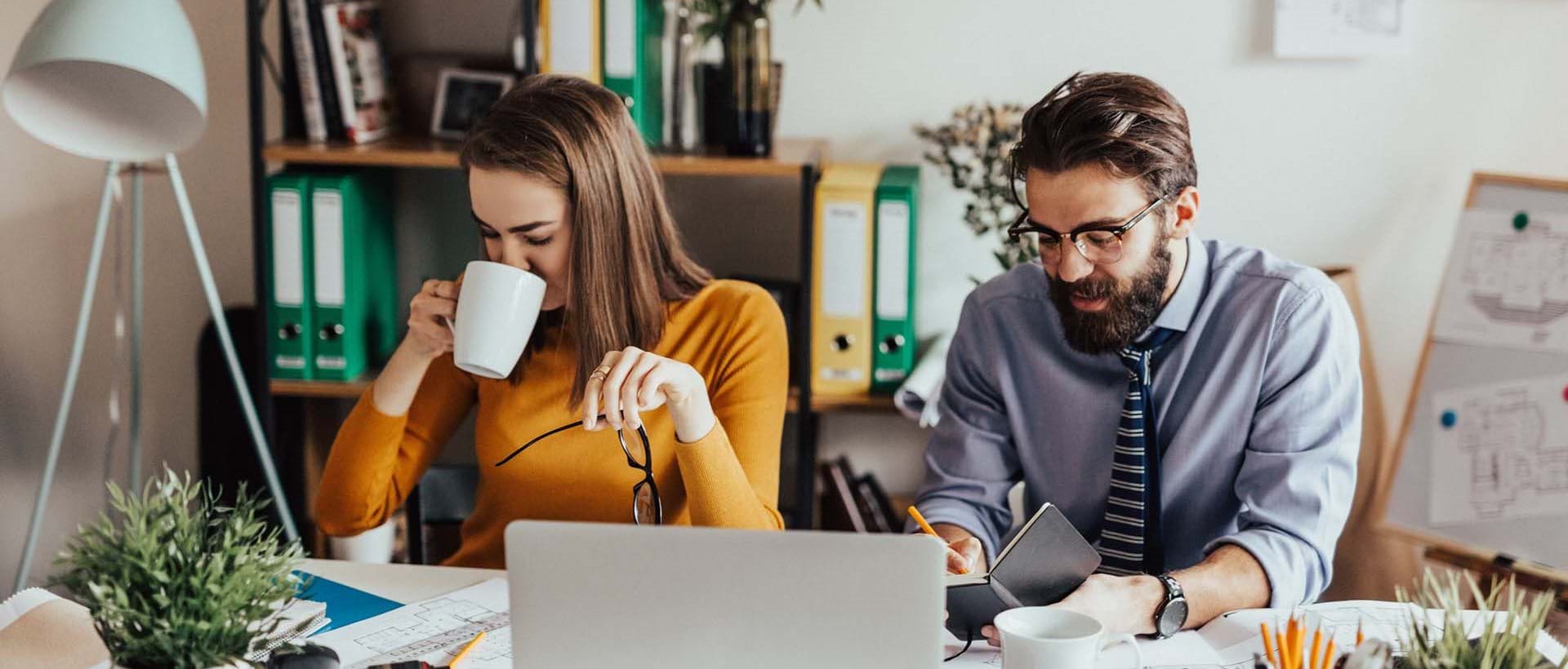 kvinna och man vid skrivbord använder dator och dricker kaffe